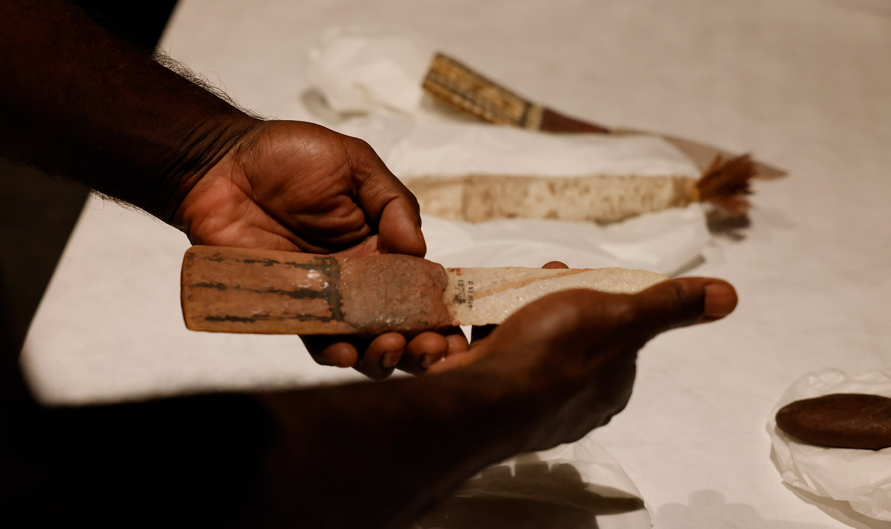 A stone knife being held up and various artefacts on a table in the background. 