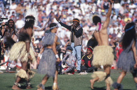 A man singing in front of a large crowd with dancers